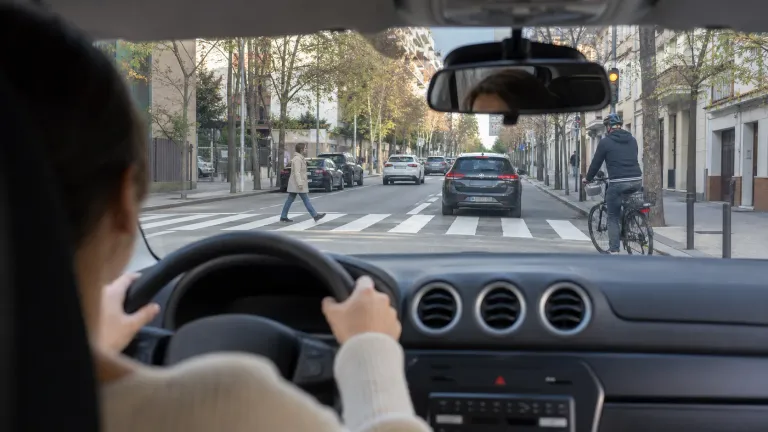 Coche de autoescuela circulando en ciudad mientras un alumno anticipa riesgos de peatones y tráfico